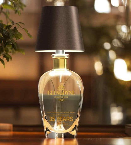 Lampa med mörkbrun skärm på en dekorativ glaskaraff med guldfärgad hals, i varm och dämpad belysning. 5
