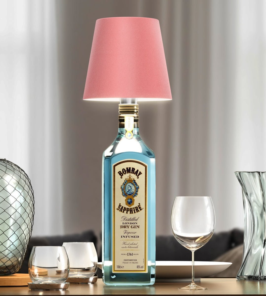 Lampa med rosa skärm på en blå ginflaska, omgiven av glas och en glasvas på ett bord. 4