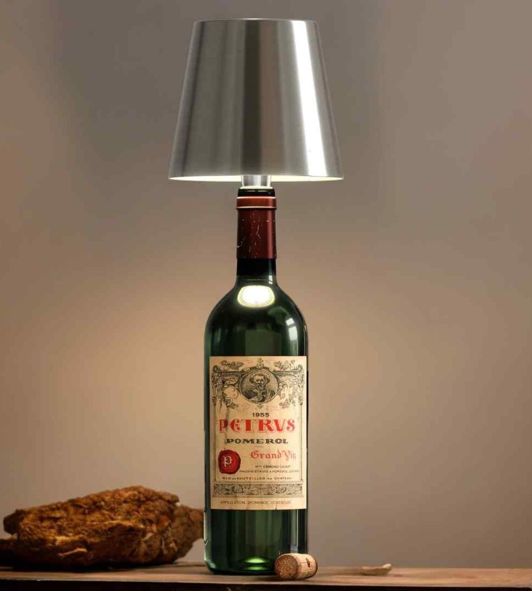 Lampa med silverfärgad metallskärm på en grön vinflaska med gammaldags etikett, bredvid en kork och en sten. 7