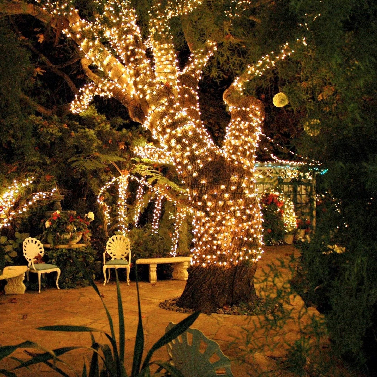 Beleuchteter Baum im romantischen Garten mit Lichterketten und weißen Gartenstühlen.