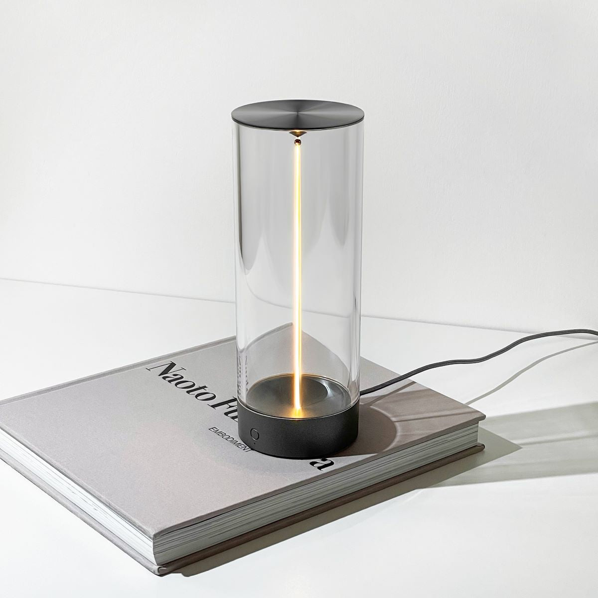 Moderne Tischlampe aus Glas und Metall, zylindrisch, auf Buch, minimalistisches Design.