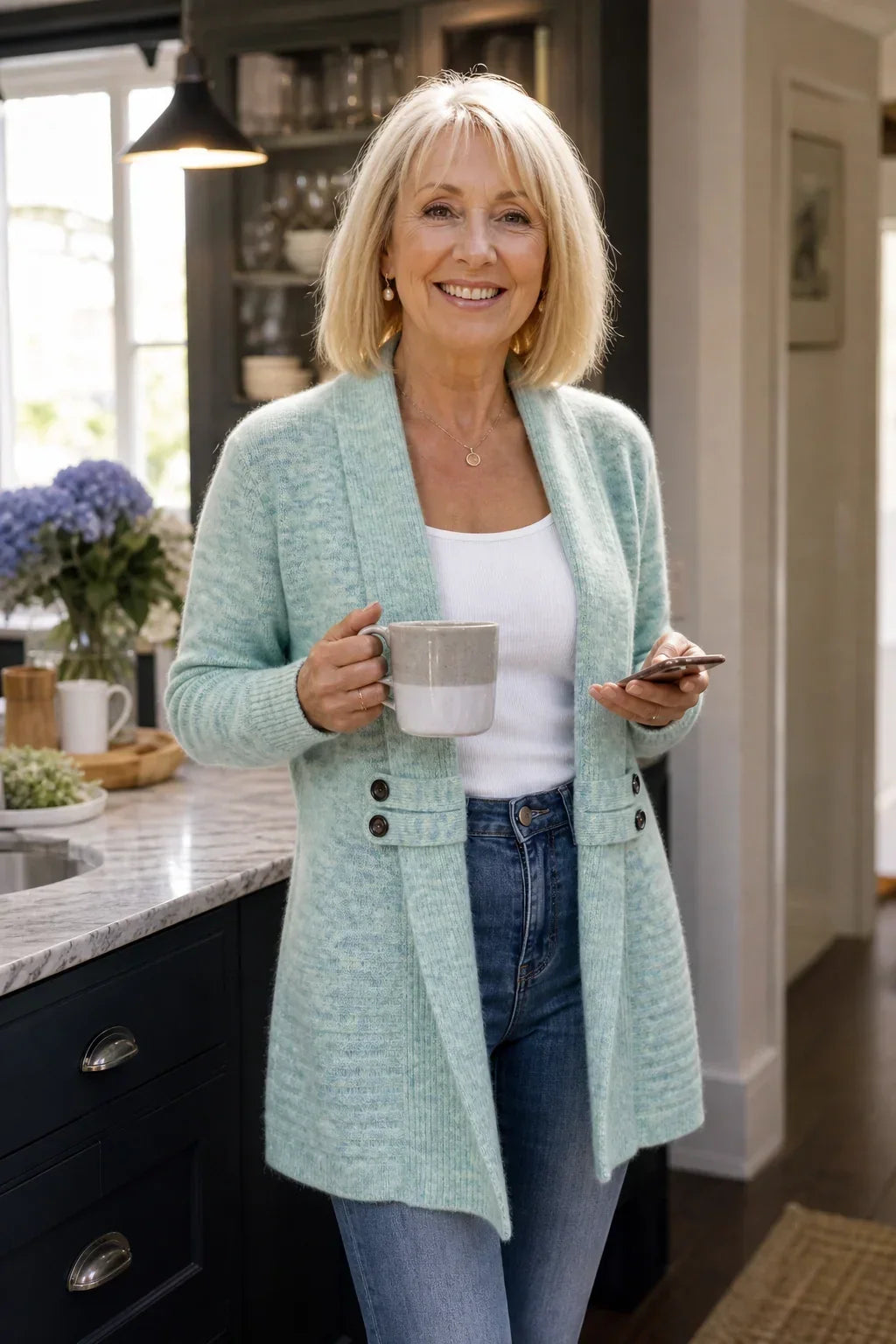 Leende kvinna i köket bär ljusturkos kofta med vit topp och jeans, kaffekopp i handen 4
