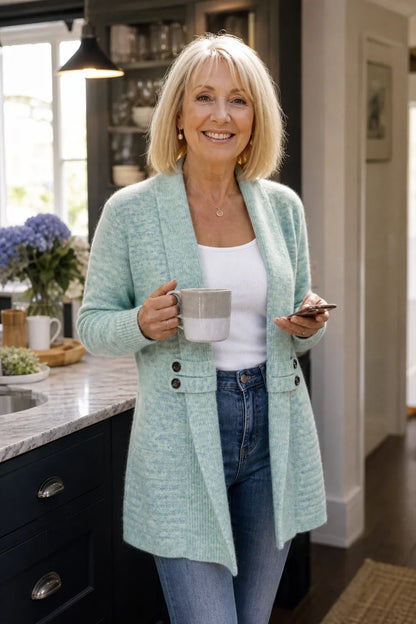 Leende kvinna i köket bär ljusturkos kofta med vit topp och jeans, kaffekopp i handen 4