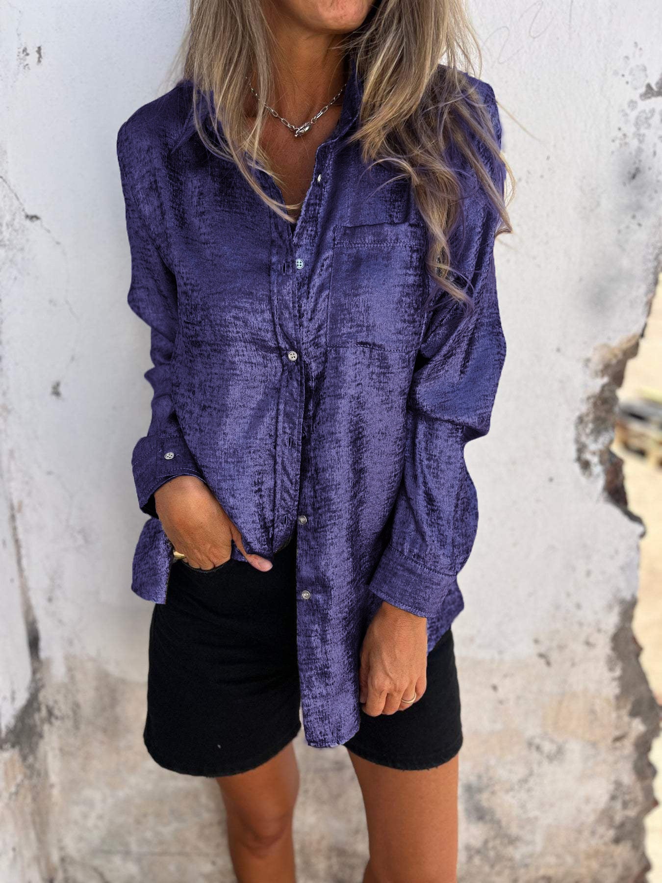 Frau in lila Satinbluse und schwarzer Shorts vor weißer Wand.