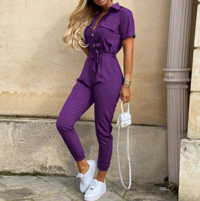 Frau in lila Jumpsuit mit weißer Handtasche und Sneakers, modisch, urbaner Stil.