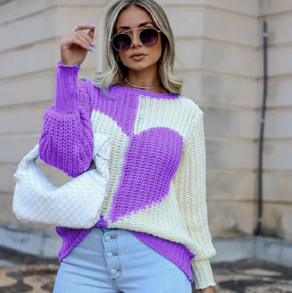 Frau mit lila-weißem Strickpullover, Herzmotiv, Sonnenbrille, weiße Tasche, Jeans.