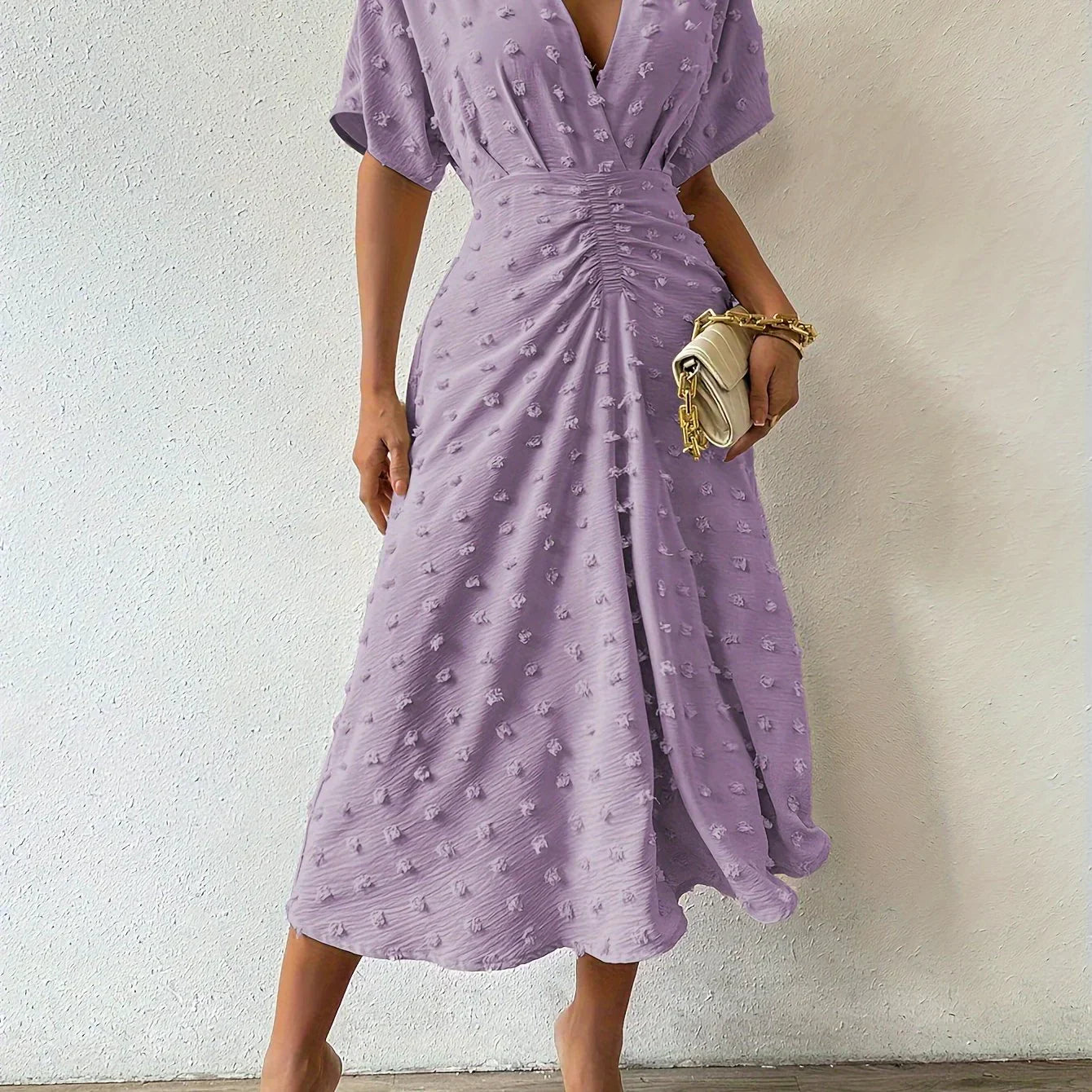 Frau in lila Sommerkleid mit V-Ausschnitt, Raffung, hält beigen Clutch, modisch elegant.