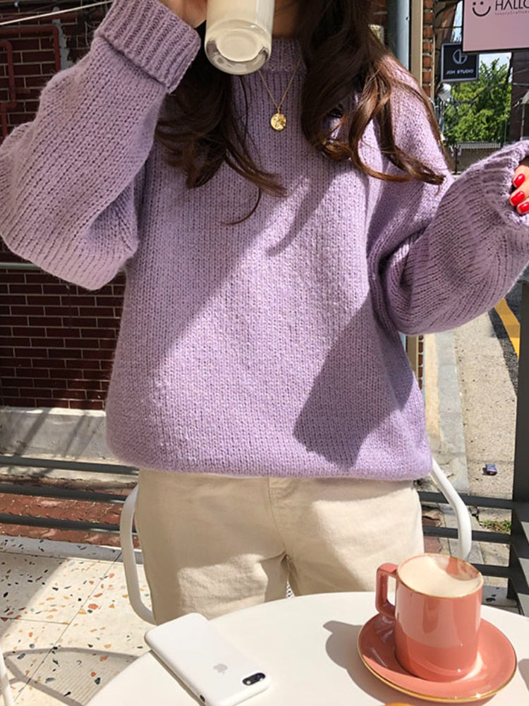 Frau in lila Strickpullover trinkt Kaffee, sitzt draußen, Smartphone und Tasse auf Tisch.