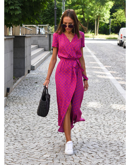 Frau im pinken Wickelkleid mit Punkten, trägt schwarze Tasche, weiße Sneaker, Sonnenbrille.