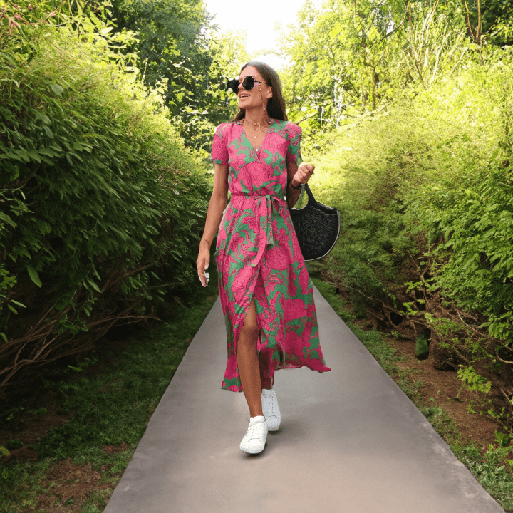 Frau in pink-grünem Sommerkleid, Sonnenbrille, weiße Sneaker, schwarze Tasche, Gartenweg.