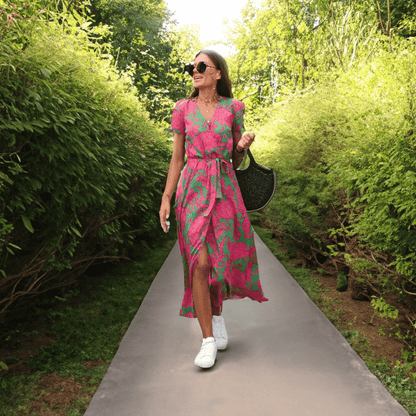 Frau in pink-grünem Sommerkleid, Sonnenbrille, weiße Sneaker, schwarze Tasche, Gartenweg.