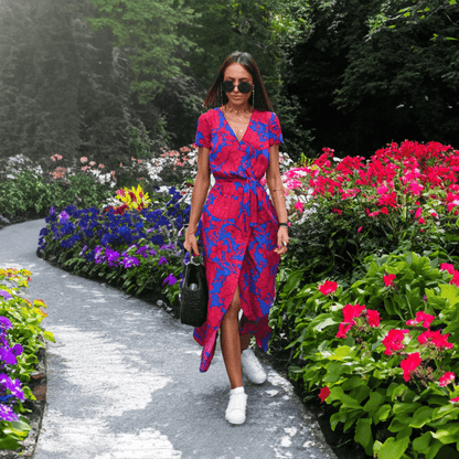 Frau in rotem Sommerkleid mit blauem Muster spaziert im blühenden Garten, trägt Sonnenbrille.
