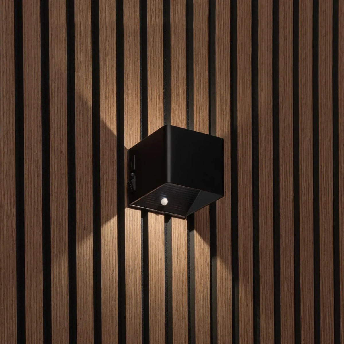Svart kubformad vägglampa på träpanel, sedd snett från sidan med strömbrytare och rörelsesensor synliga 1