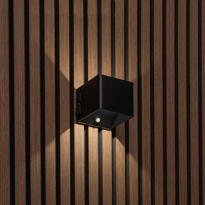 Svart kubformad vägglampa på träpanel, sedd snett från sidan med strömbrytare och rörelsesensor synliga 1