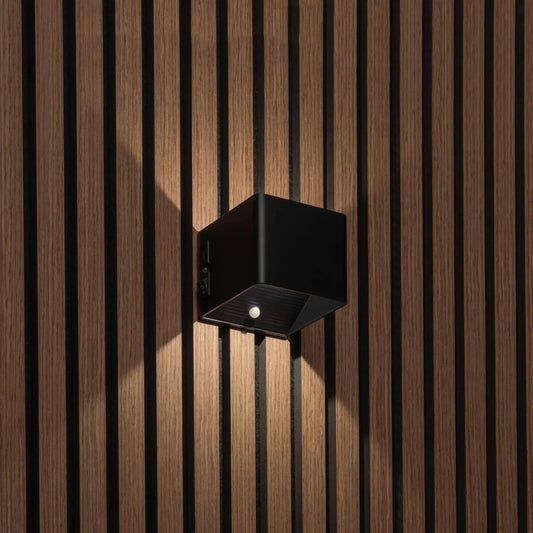 Svart kubformad vägglampa på träpanel, sedd snett från sidan med strömbrytare och rörelsesensor synliga 1