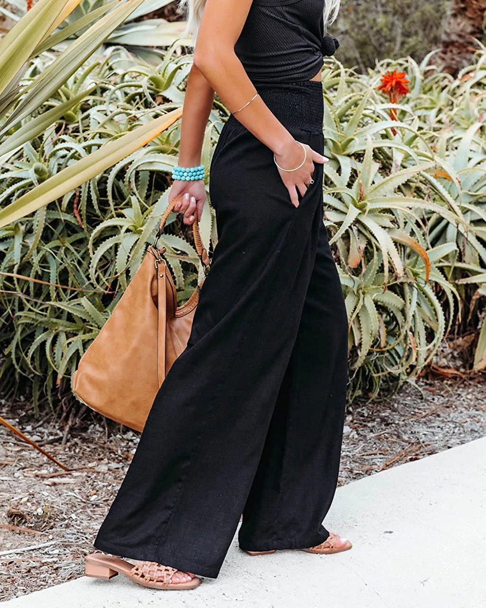 Frau in schwarzer Hose, braune Ledertasche, Sommermode, lässiger Stil, Gartenhintergrund.