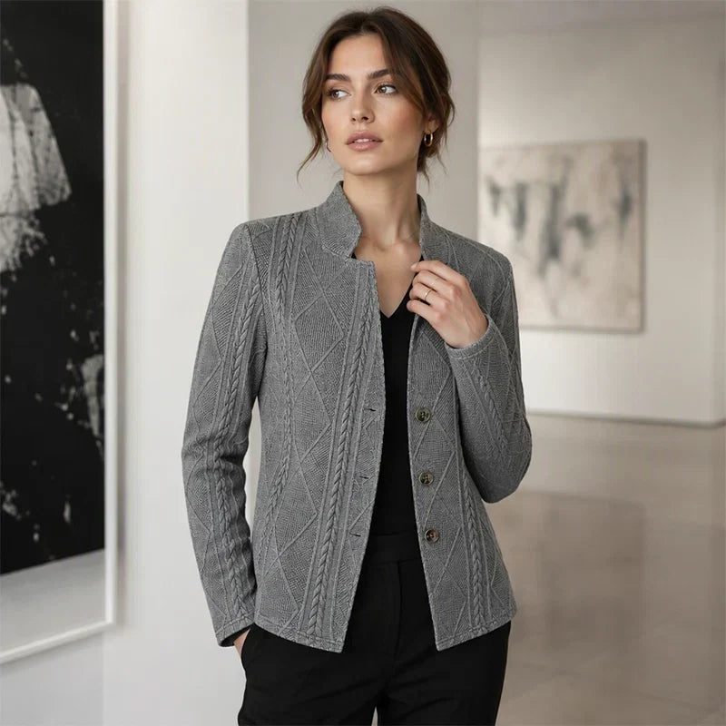 Grå strukturerad blazer bärd över svart topp av en kvinna som står i ett ljust galleri med handen i fickan. 11