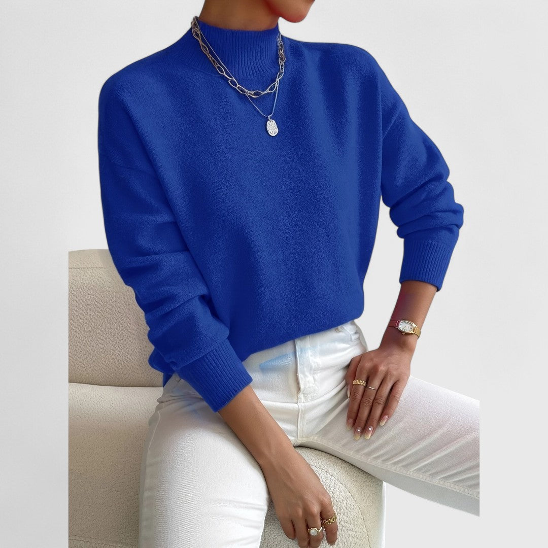 Modell i klarblå polotröja med ribbad krage och muddar, vita jeans och silverfärgade halsband, sittande på en vit fåtölj. 11