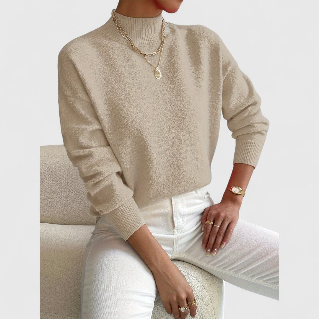 Modell i beige polotröja med ribbad krage och muddar, vita jeans och guldfärgade halsband, sittande på en vit fåtölj. 7