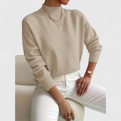 Modell i beige polotröja med ribbad krage och muddar, vita jeans och guldfärgade halsband, sittande på en vit fåtölj. 7