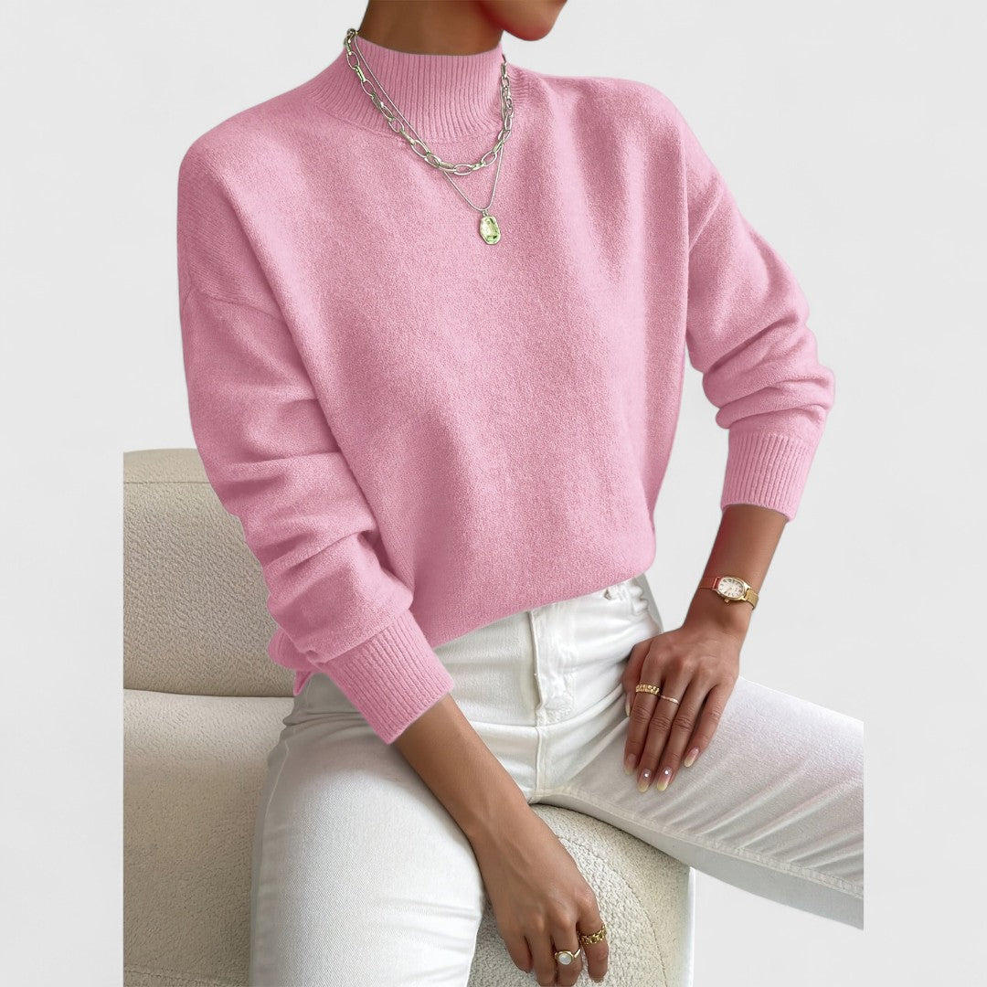 Modell i rosa polotröja med ribbad krage och muddar, vita jeans och silverfärgade halsband, sittande på en vit fåtölj. 5