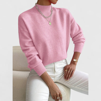 Modell i rosa polotröja med ribbad krage och muddar, vita jeans och silverfärgade halsband, sittande på en vit fåtölj. 5