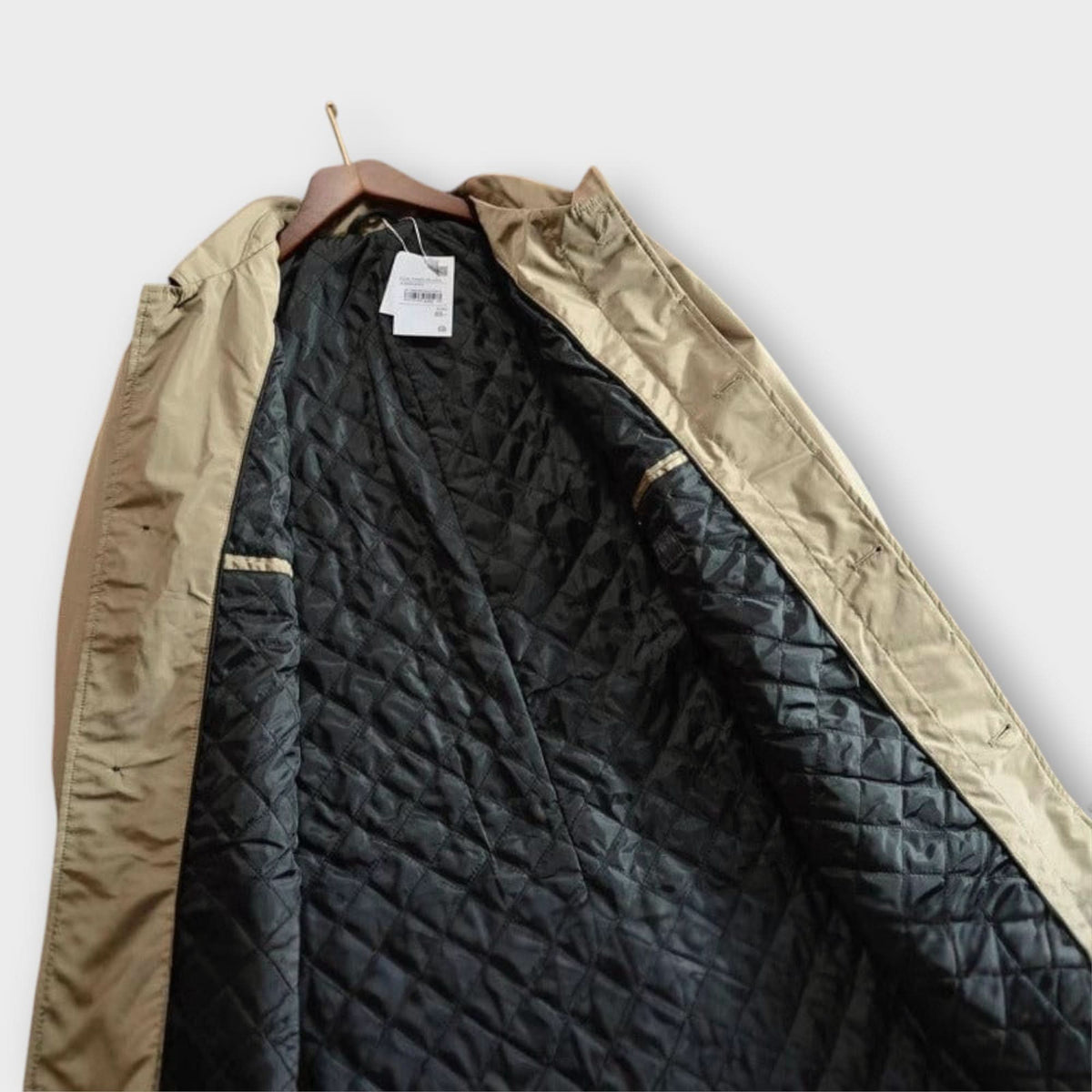 Insidan av den beige kappan på galge, med svart rutsydd diamantquiltad fodring och en hängande prislapp. 6