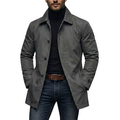 Man bär den grå kappan öppen över svart polotröja och jeans, med händerna i fickorna, sedd framifrån. 1