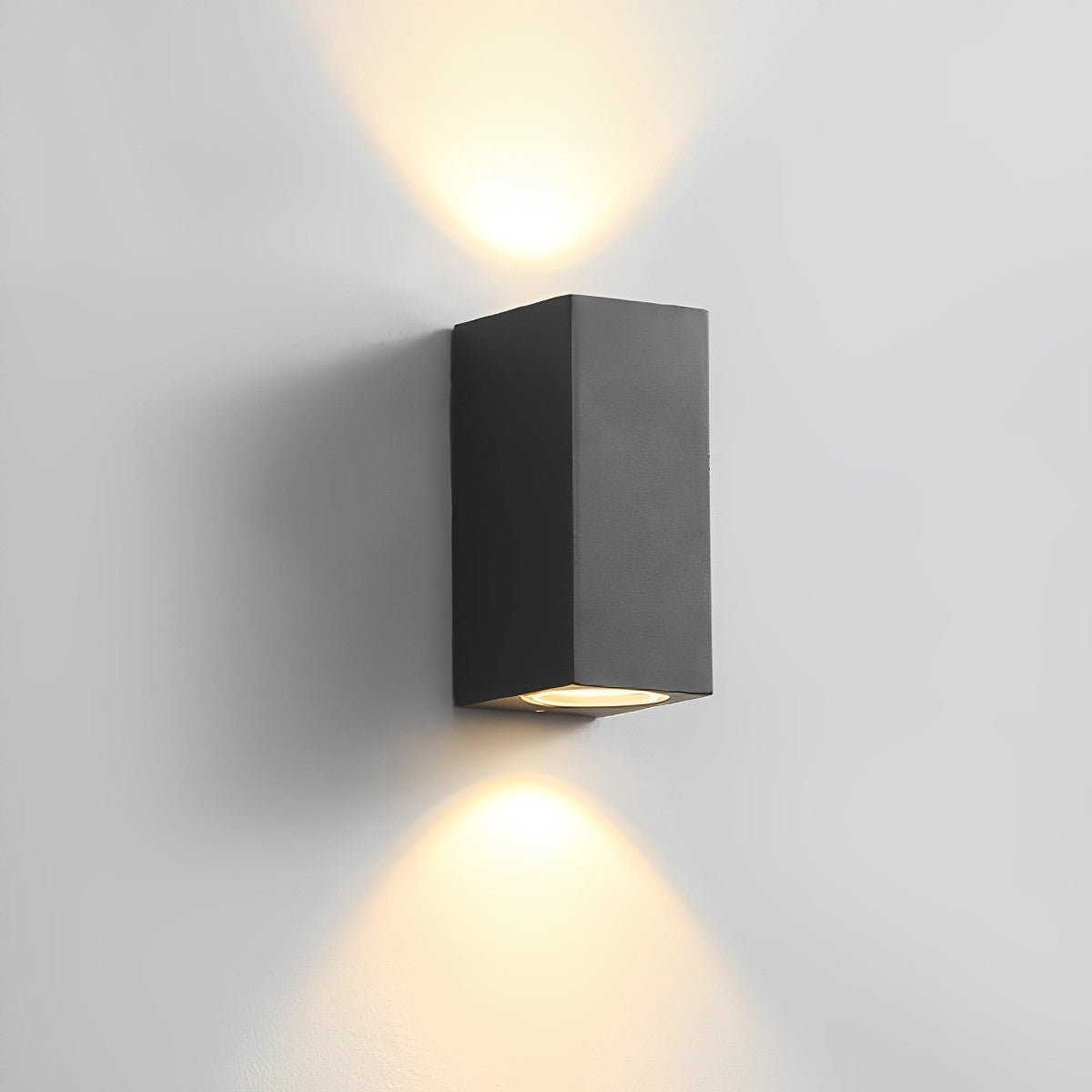 Svart rektangulär vägglampa på ljusgrå vägg som sprider varmt ljus uppåt och nedåt 12