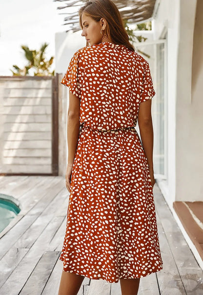 Frau in rotem Sommerkleid mit weißen Punkten, steht auf Holzterrasse, seitlich zur Kamera.