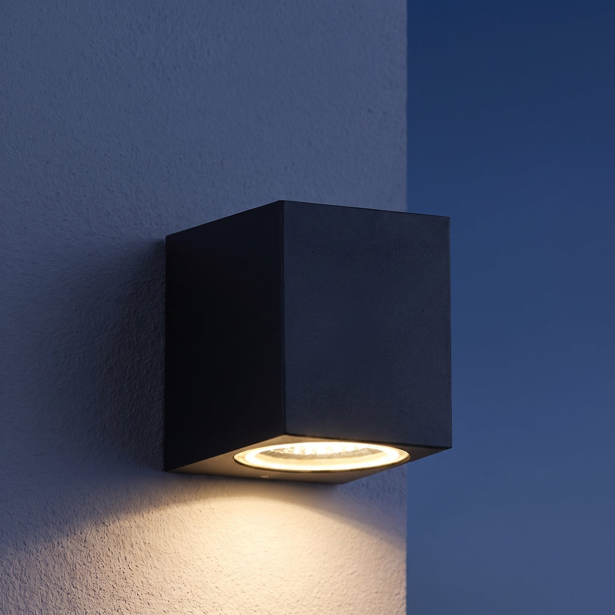 Svart kubformad vägglampa på texturerad vägg i skymning, med varmt ljus nedåt 2