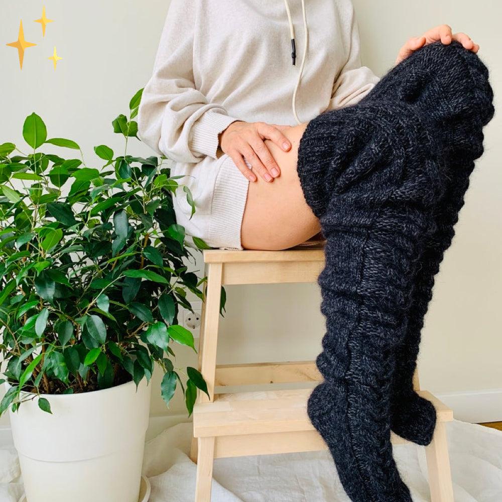 Frau in cremefarbenem Hoodie mit schwarzen Strick-Overknee-Socken auf Holzleiter neben Pflanze.