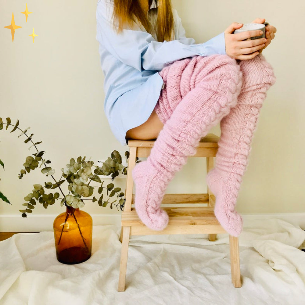 Frau in blauer Bluse trägt rosa gestrickte Overknee-Socken, sitzt auf Holzstuhl, hält Tasse.