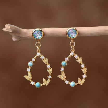 Goldene Ohrringe mit Opal, Zirkonia und Schmetterlingsdesign, hängend an Ast.