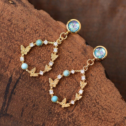 Goldene Ohrringe mit Opal, Zirkonia und Schmetterlingsdesign auf Holzoberfläche.