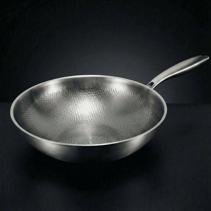 Wokpannan snett från sidan mot mörk bakgrund, med skinande hamrat mönster på insidan och ett långt, matt metallhandtag. 1