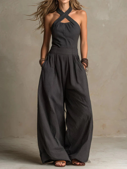Kvinna i mörkgrå, nålstrimmig jumpsuit med korsade halsband, hög midja och mycket vida ben, händerna i fickorna, framifrån. 1
