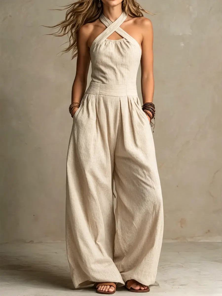 Kvinna i beige jumpsuit med korsade halsband, markerad midja och vida ben, händerna i fickorna, fotograferad framifrån mot beige vägg. 2