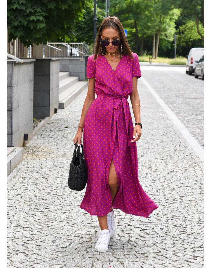 Frau in pinkem Wickelkleid mit Punkten, trägt schwarze Tasche, weiße Sneaker, Sonnenbrille.