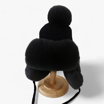 Schwarze Wintermütze mit Kunstfell, Bommel und Ohrenschützern auf Holzständer.