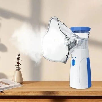 Vit och blå handhållen nebulisator med ansiktsmask som avger fin dimma, stående på ett träbord bredvid en bok och en liten växt. 1