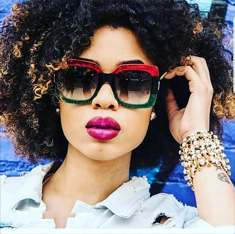 Frau mit Afro, trägt große, bunte Sonnenbrille und goldene Armbänder vor blauer Wand.