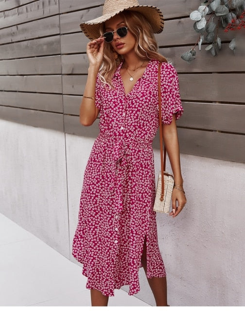 Frau im roten Sommerkleid mit Blumenmuster, Strohhut und Sonnenbrille vor Holzwand.