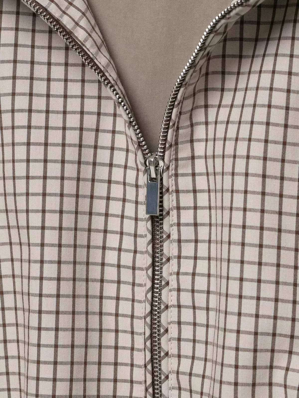 Detaljbild av jackans dragkedja i silverfärg med skjutare mot rutigt tyg i beige och brunt. 6