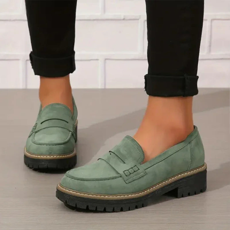 Gröna mockaliknande loafers med grov svart sula, burna av en modell i uppvikta svarta byxor mot vit tegelvägg. 1