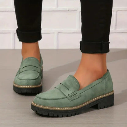 Gröna mockaliknande loafers med grov svart sula, burna av en modell i uppvikta svarta byxor mot vit tegelvägg. 1