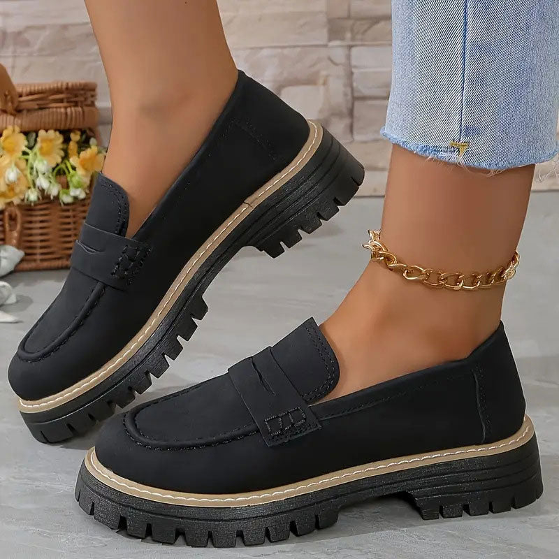Svarta loafers med tjock plattformssula och beige sulkant, burna med guldig fotkedja framför en korg med blommor. 12