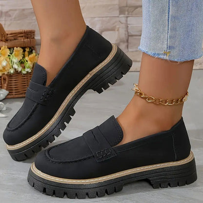 Svarta loafers med tjock plattformssula och beige sulkant, burna med guldig fotkedja framför en korg med blommor. 12