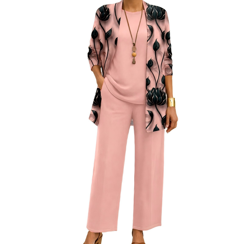 Modell i rosa set med lång kofta med svarta blommotiv, matchande topp och vida byxor samt halsband. 2