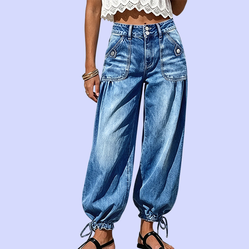 Närbild framifrån på blå jeans med hög midja, knäppta framfickor och knytband vid fållen mot lila bakgrund. 10